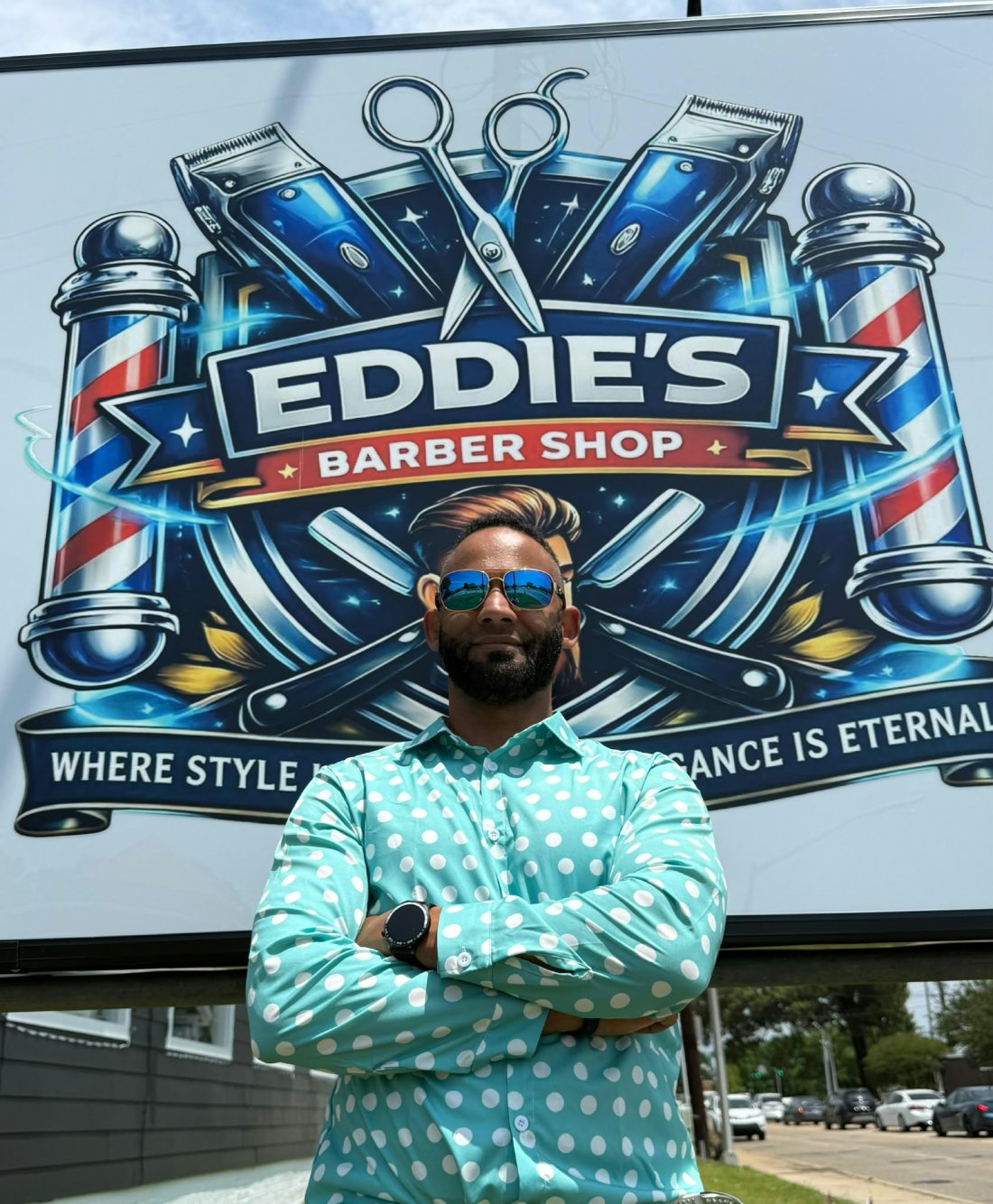 Eddie standing with his arms crossed in a mint-green polka-dot shirt and mirrored sunglasses, in front of the shop's illuminated billboard showing the Eddie's Barber Shop crest.