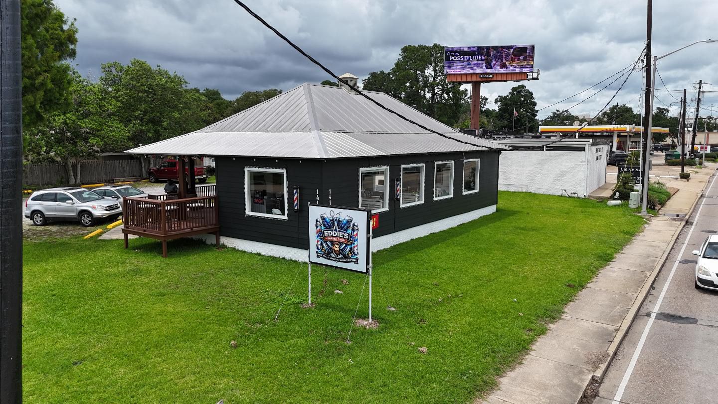 Eddie's Barber Shop from the street: a black board-and-batten building with a silver tin roof, white-trimmed windows, a wooden front deck, and a yard sign bearing the shop's crest.