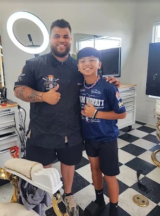 Adahias and a young client giving a thumbs-up together after a fresh cut, standing on the checkerboard floor.