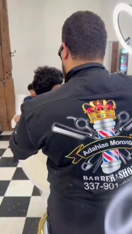 Adahias mid-cut in his embroidered barber shirt, working on a client beside the shop's checkerboard floor.